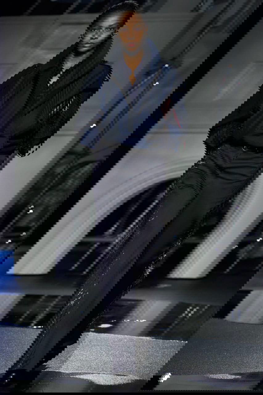 Модель в укороченной синей куртке и боком виднеется лужа на полу подиума; в фоне архитектурная декорация и подсветка шоу.