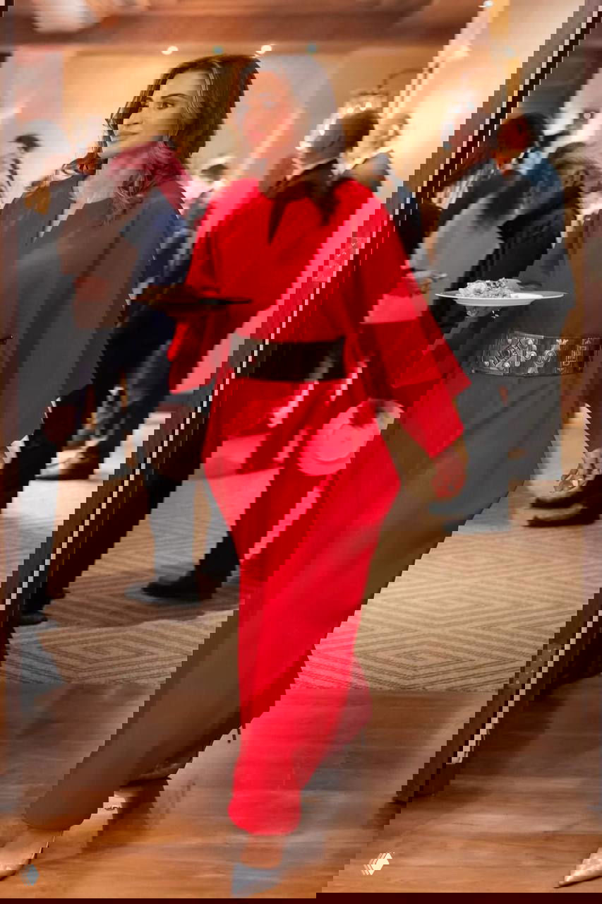 Женщина в длинном пальто-пончо серого оттенка идёт по красной дорожке рядом с гостем; официальный выход на открытой площадке при ярком дневном освещении.