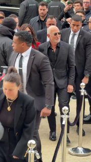 Мужчина в солнечных очках и костюме проходит мимо ограждений с охраной; на заднем плане толпа и фотографы у входа на показ.