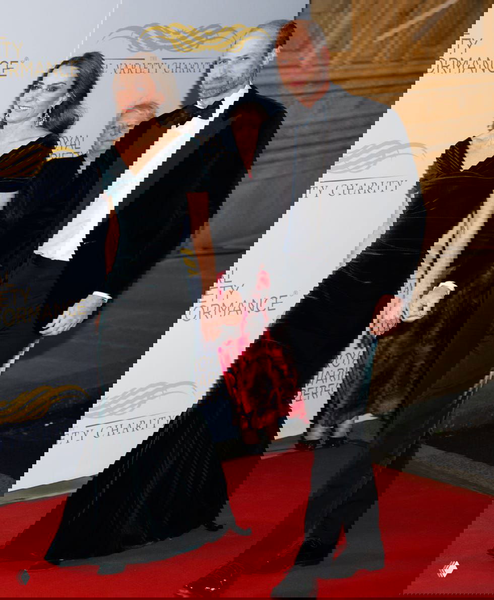 Пара на красной дорожке: женщина в тёмно‑зелёном платье и мужчина в чёрном смокинге, общий план у входа.