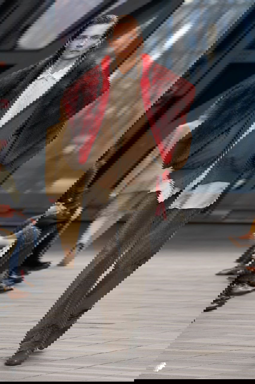 Модель в коричневой бомбер-короткой куртке с красным меховым накидом и брюками, образ сочетает мужские и женские элементы в минималистичной гамме.