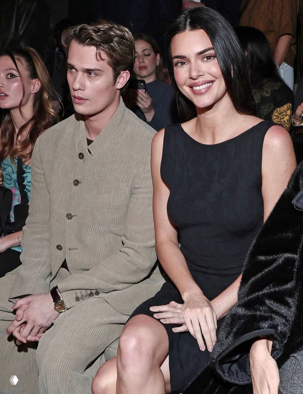 Кендалл Дженнер и Николас Галицин сидят в первом ряду модного показа, зрители и камеры видны в фоне — атмосферный кадр с шоу.