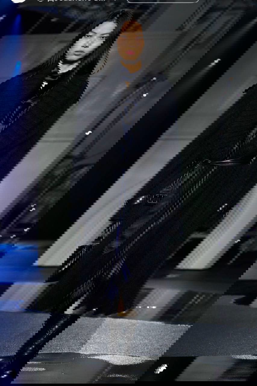 Модель в тёмном длинном пальто и шелковистой рубашке; подиум с отражениями и строгой архитектурой задника шоу Burberry.