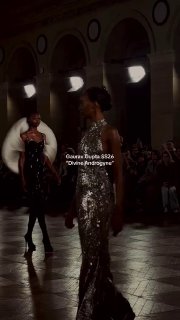 Блестящее тёмное платье на подиуме Gaurav Gupta: модели и дизайнер присутствуют в кадре, публика наблюдает показ в атмосферном интерьере.