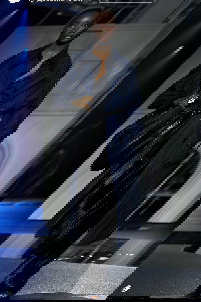 Модель в синем плиссированном тренче и широких бархатных брюках шагает по подиуму с отражающим покрытием и мостовой декорацией.