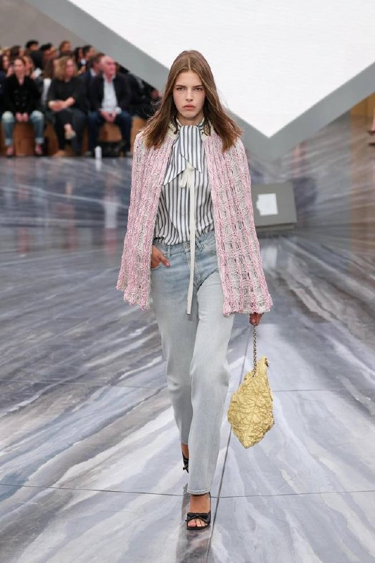 Модель в нежно‑розовой накидке Chloé и светлых джинсах идёт по подиуму; образ демонстрирует текстуру, прозрачность и пастельную палитру сезона.