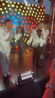 Исполнители в белых костюмах на фоне цветных огней и видеодекора, общая композиция кадра подчёркивает праздничный настрой шоу.