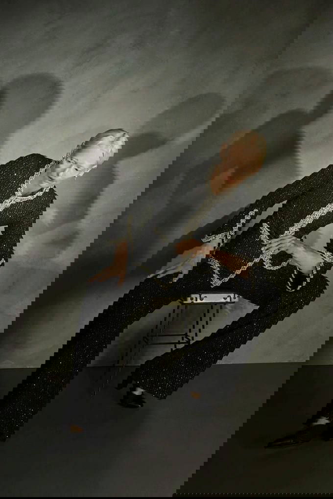 Чёрно‑белое фото: модель сидит на табурете в твидовом жакете и широких брюках Chanel; текстура ткани и контрастная отделка видны в кадре.