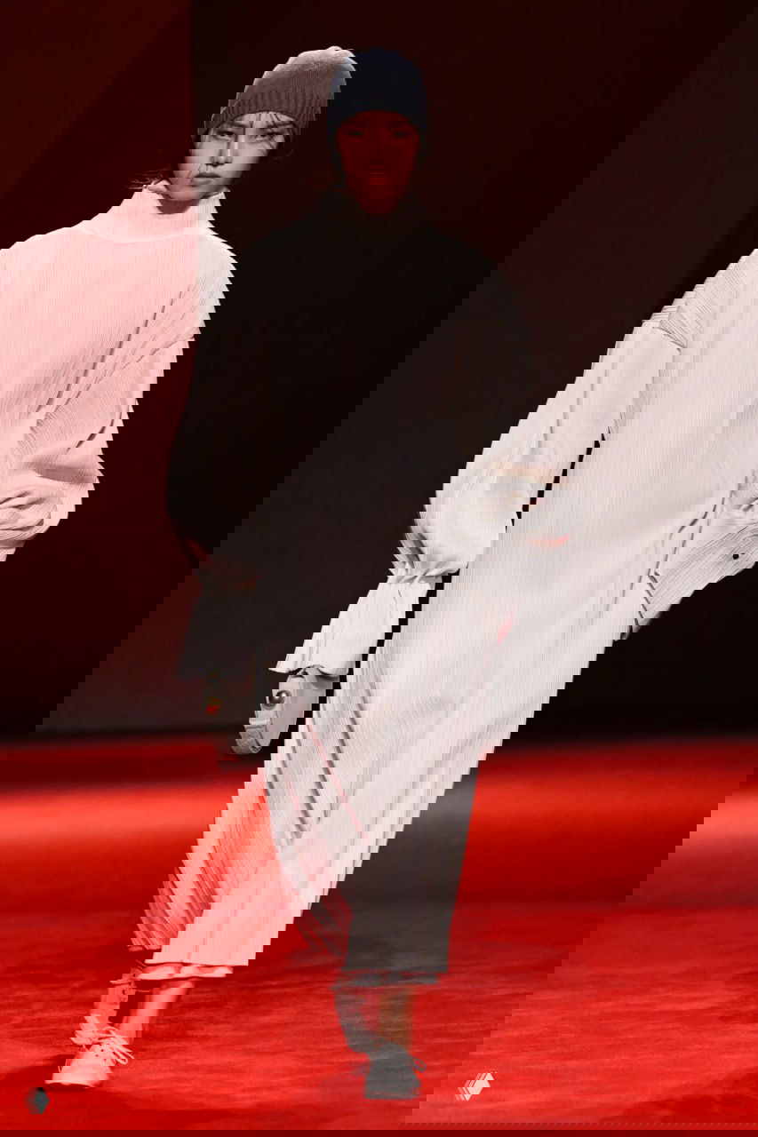 Модель в длинном свободном белом платье с высоким горлом и вязаной шапке идёт по подиуму — в коллекции виден минимализм форм и комфортный силуэт.