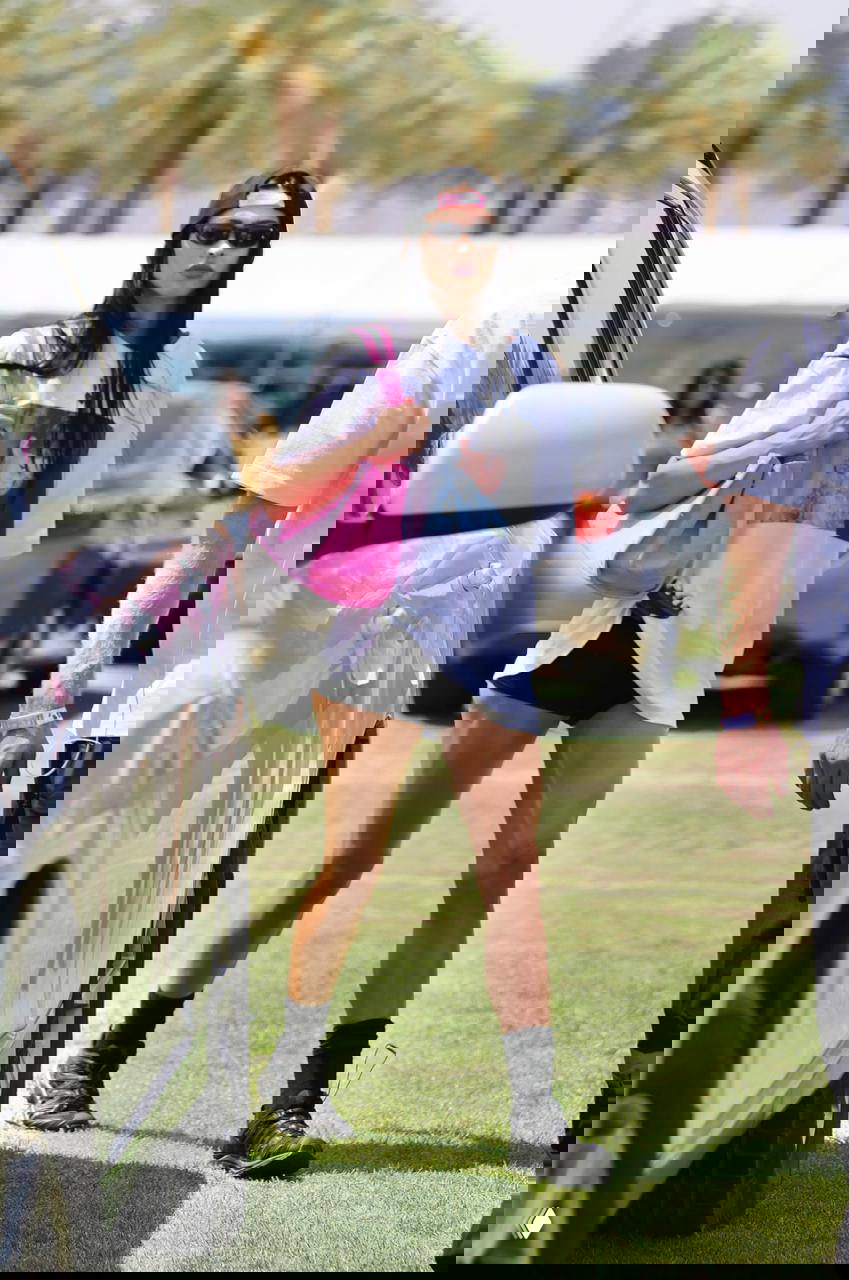 Человек в большой футболке и розовой сумке идёт по парковке фестиваля между автомобилями, на заднем плане пальмы и яркий солнечный день.