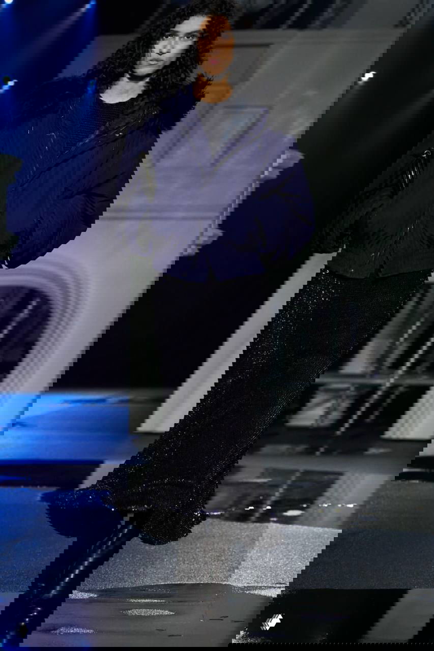 Модель в тёмно‑синей стёганой куртке с меховым воротником и блестящим платьем; подиум имитирует ночной Лондон с лужами.