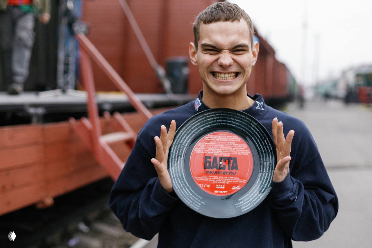 Молодой человек улыбается и держит пластинку с логотипом «Баста» на фоне товарных вагонов — реплика поезда, воссоздающая атмосферу туров конца 90-х.