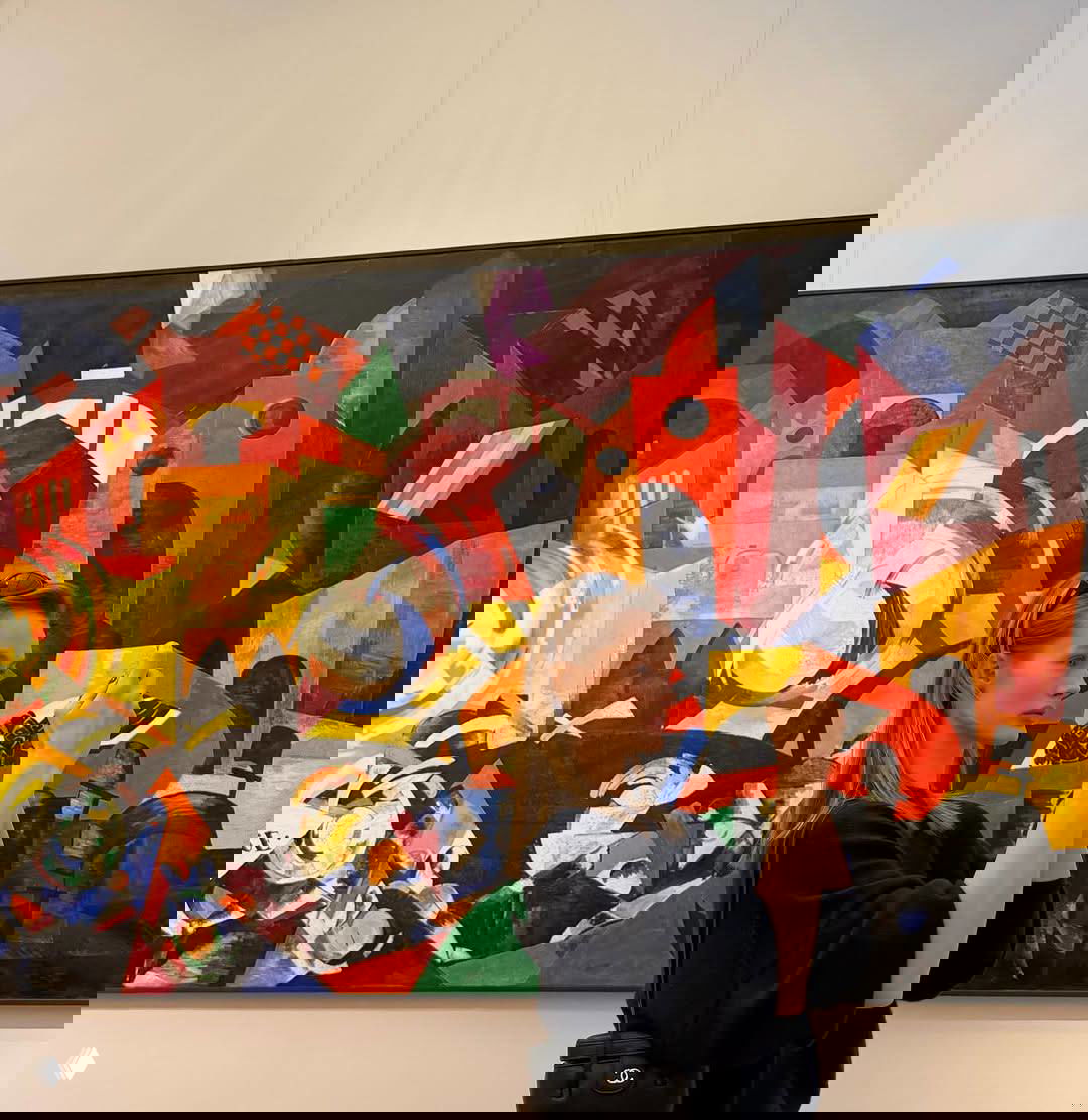 Модель фотографирует на смартфон большое яркое полотно авангардного направления с геометрическими и цветными фрагментами в экспозиции.