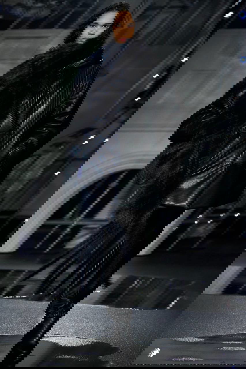 Модель в короткой тёмной куртке поверх длинного блестящего платья; отражения света на мокром подиуме создают драматичный эффект.