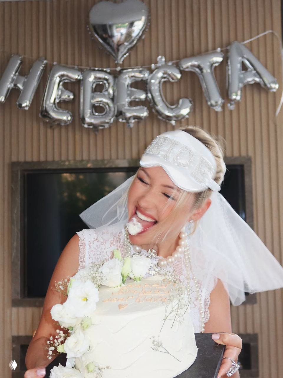 Клава смеётся и ест кусок торта в белом вуале и маске 'BRIDE'; на заднем плане видны серебристые фольгированные буквы «НЕВЕСТА».