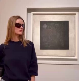 Модель в тёмной одежде стоит рядом с минималистской квадратной картиной в рамке; спокойная художественная сцена в музее.