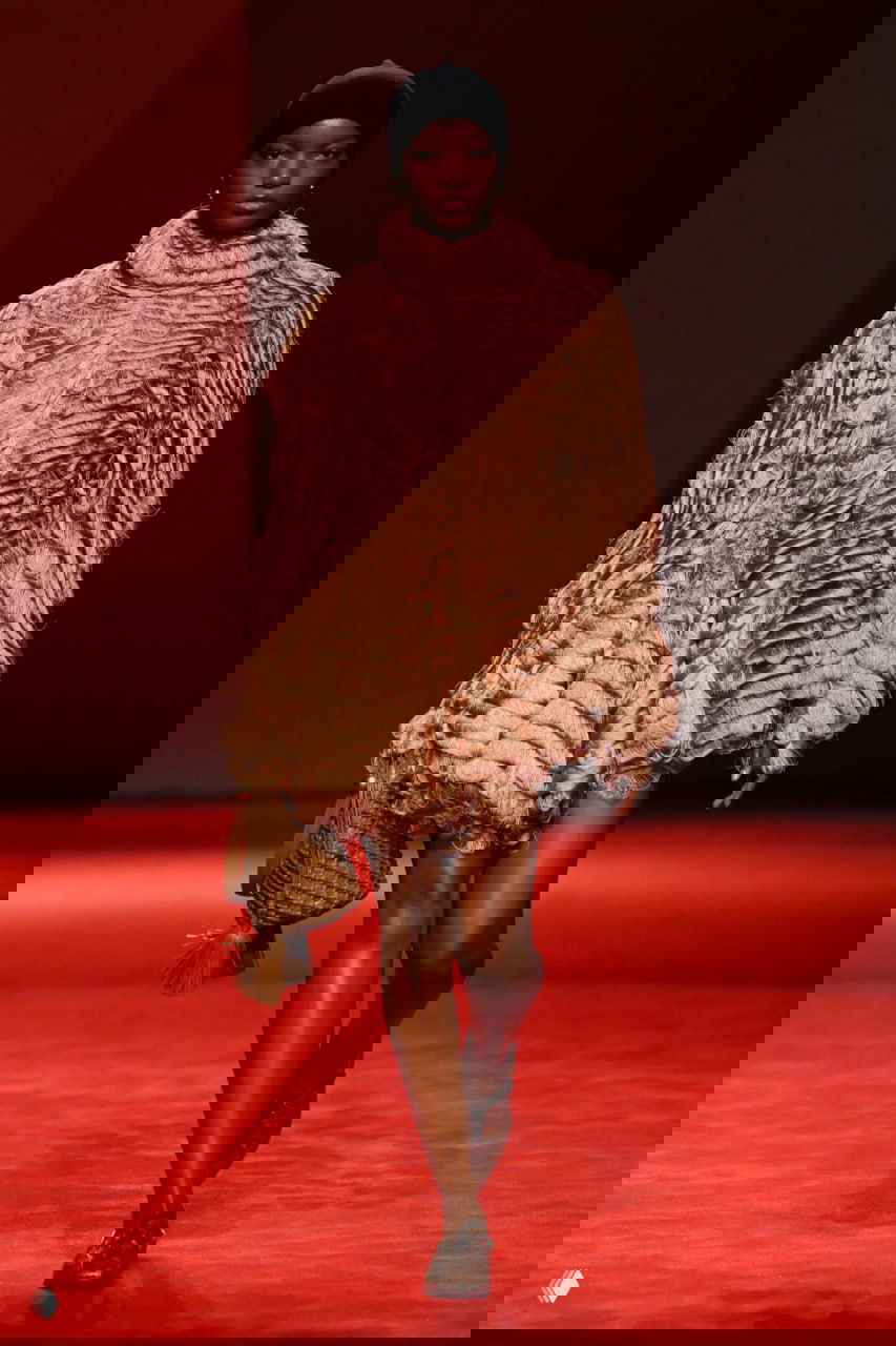 Темнокожая модель в пышной фактурной коричневой накидке несёт сумку‑плетёнку на красном подиуме — сочетание текстуры и аксессуаров в показе.