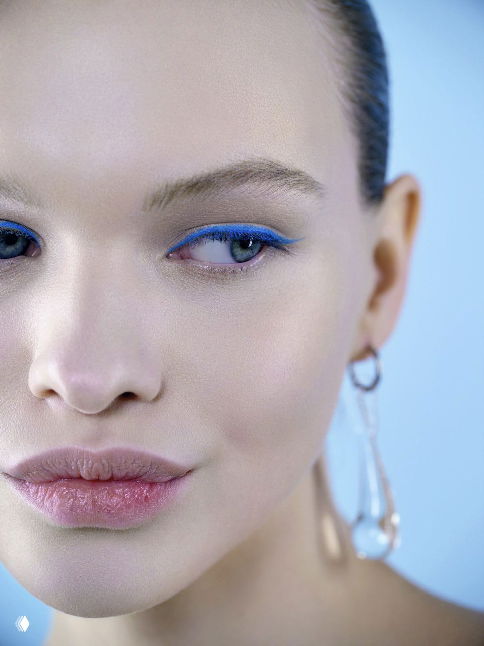 Крупный план лица с графичной ярко-синей стрелкой вдоль верхнего века, внимание на текстуре кожи, губах и серьгах в голубом фэшн-кадре.