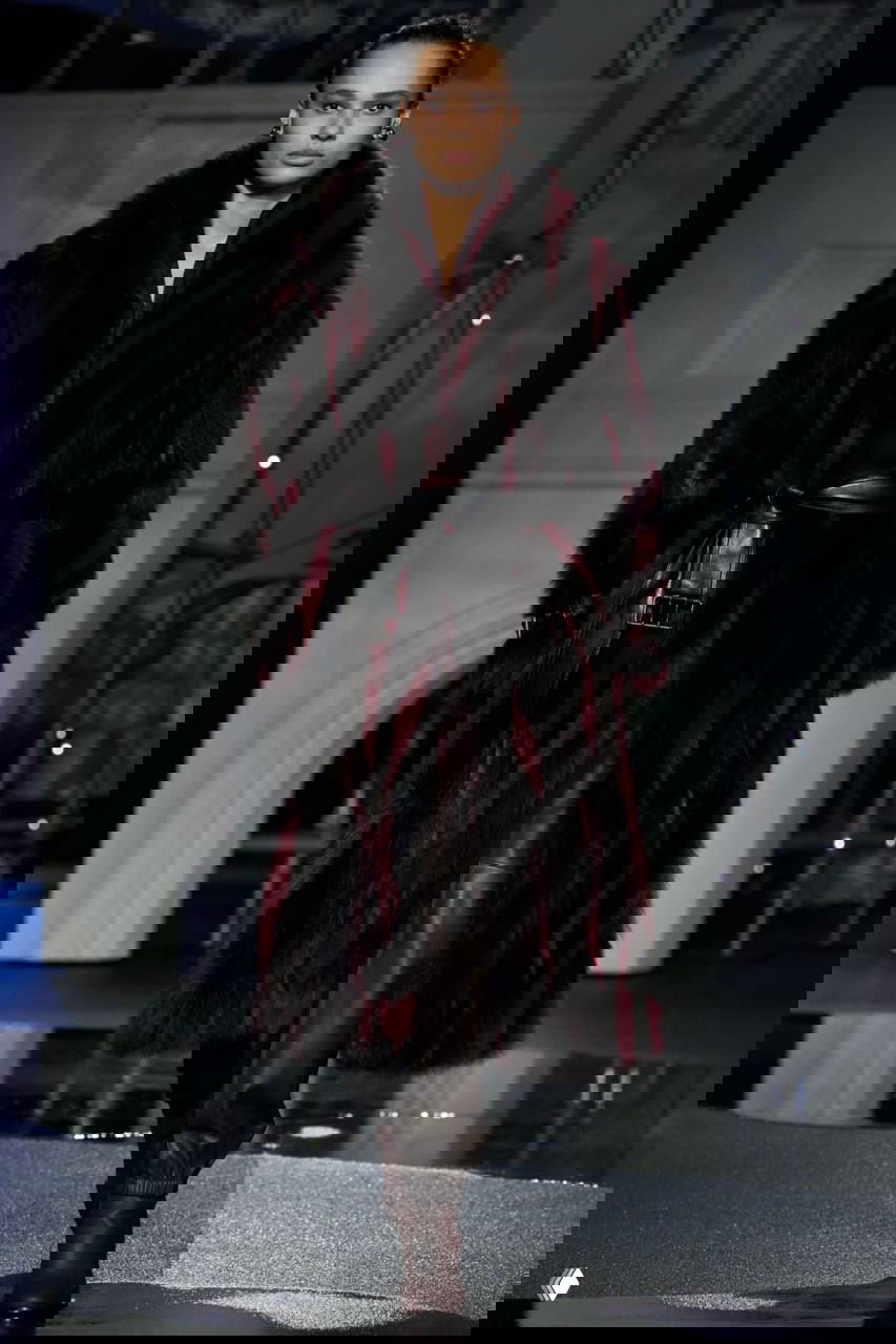 Модель в длинной шубе тёмного тона и широким поясом идёт по подиуму с зеркальными лужами и архитектурным фоном шоу Burberry.