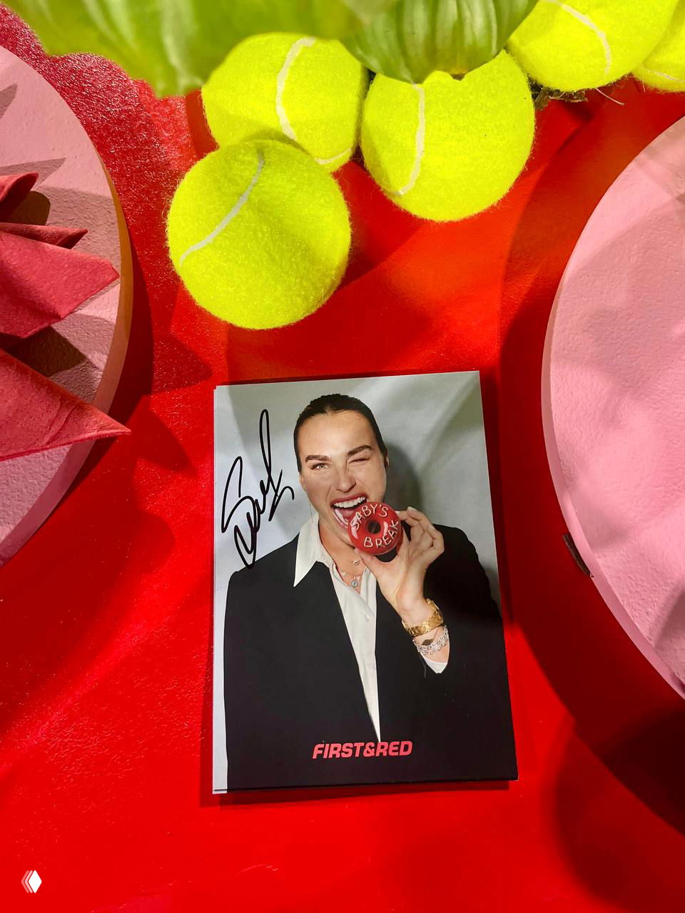 Автографированная полароидная фотография Арины Соболенко среди теннисных мячей и декора First&Red на столе мероприятия.