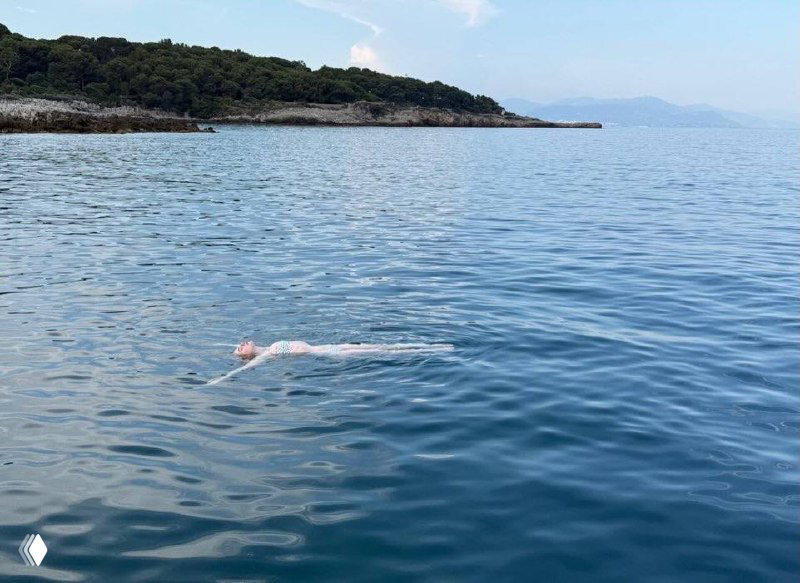 Женщина плавает на спине в открытом море; спокойная поверхность воды и береговая линия с лесистыми холмами на горизонте.