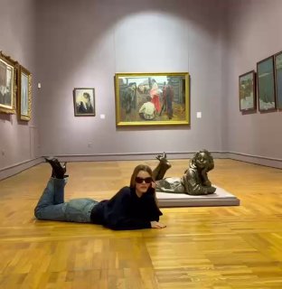 Женщина в тёмной одежде лежит на паркете музейного зала рядом с бронзовой скульптурой на фоне нескольких картин в рамах.