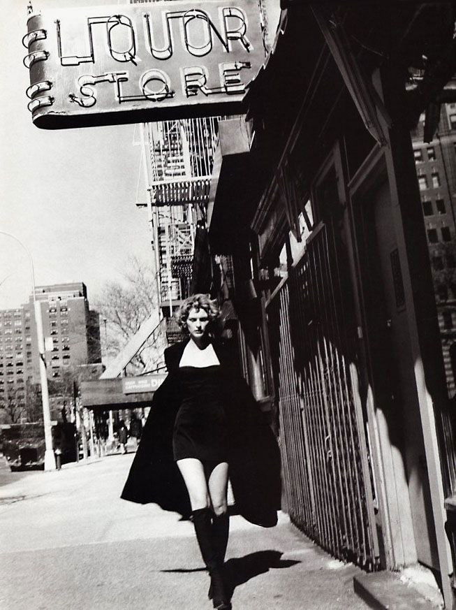 Уличный чёрно‑белый кадр: модель в коротком платье и плаще идёт мимо вывески 'LIQUOR STORE', городская винтажная сцена 1997 года.