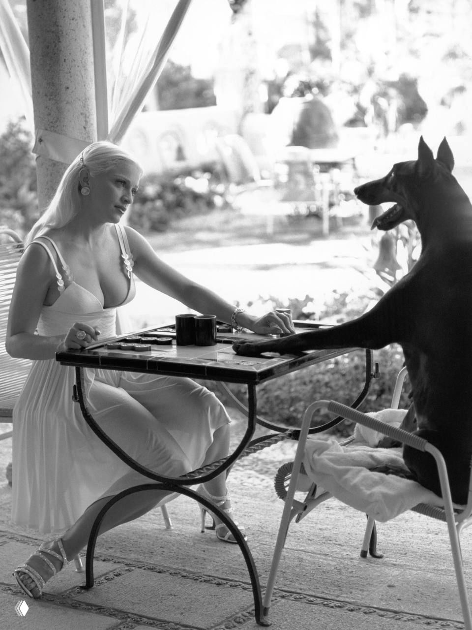 Мадонна сидит за столом на веранде и играет с собакой; чёрно‑белая сцена в элегантном платье и аксессуарах, съёмка Стивена Майзеля.