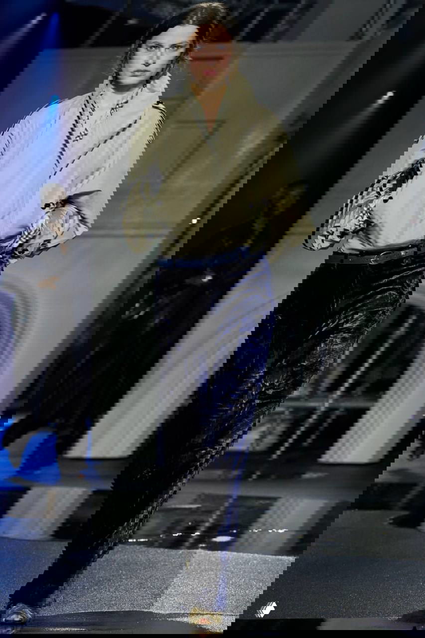 Модель в светлом тренче с фирменной клеткой на подкладке и бархатных брюках шагает по подиуму с имитацией ночного городского ландшафта.