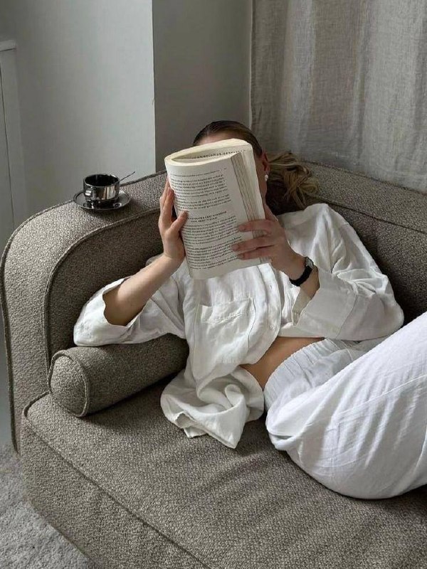 Человек, прикрыв лицо книгой, лежит на бежевом диване рядом с чашкой кофе — кадр о покое, чтении и спокойной домашней атмосфере.
