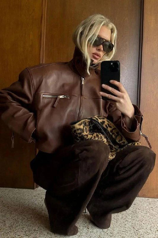 Модель в тёмно-коричневой кожаной куртке с молнией под горло и высоким воротником, сидя на корточках с леопардовой сумкой — богатая фактура и акцент на ворот.