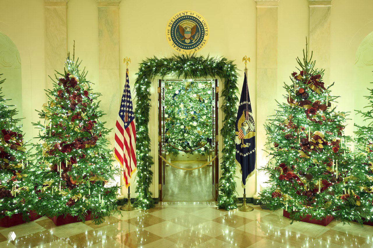 Входная арка Белого дома, окружённая нарядными ёлками и зелёными гирляндами, по бокам развеваются флаг США и президентский штандарт.