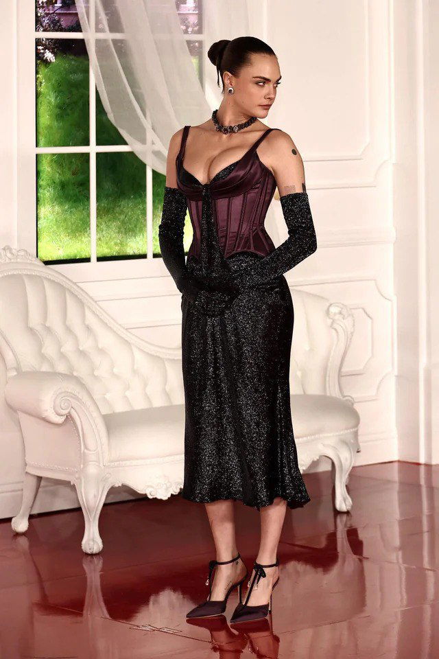 Гостья в корсете и тёмной юбке с длинными перчатками стоит у белой декоративной мебели и окна — образ в готических и винтажных мотивах.