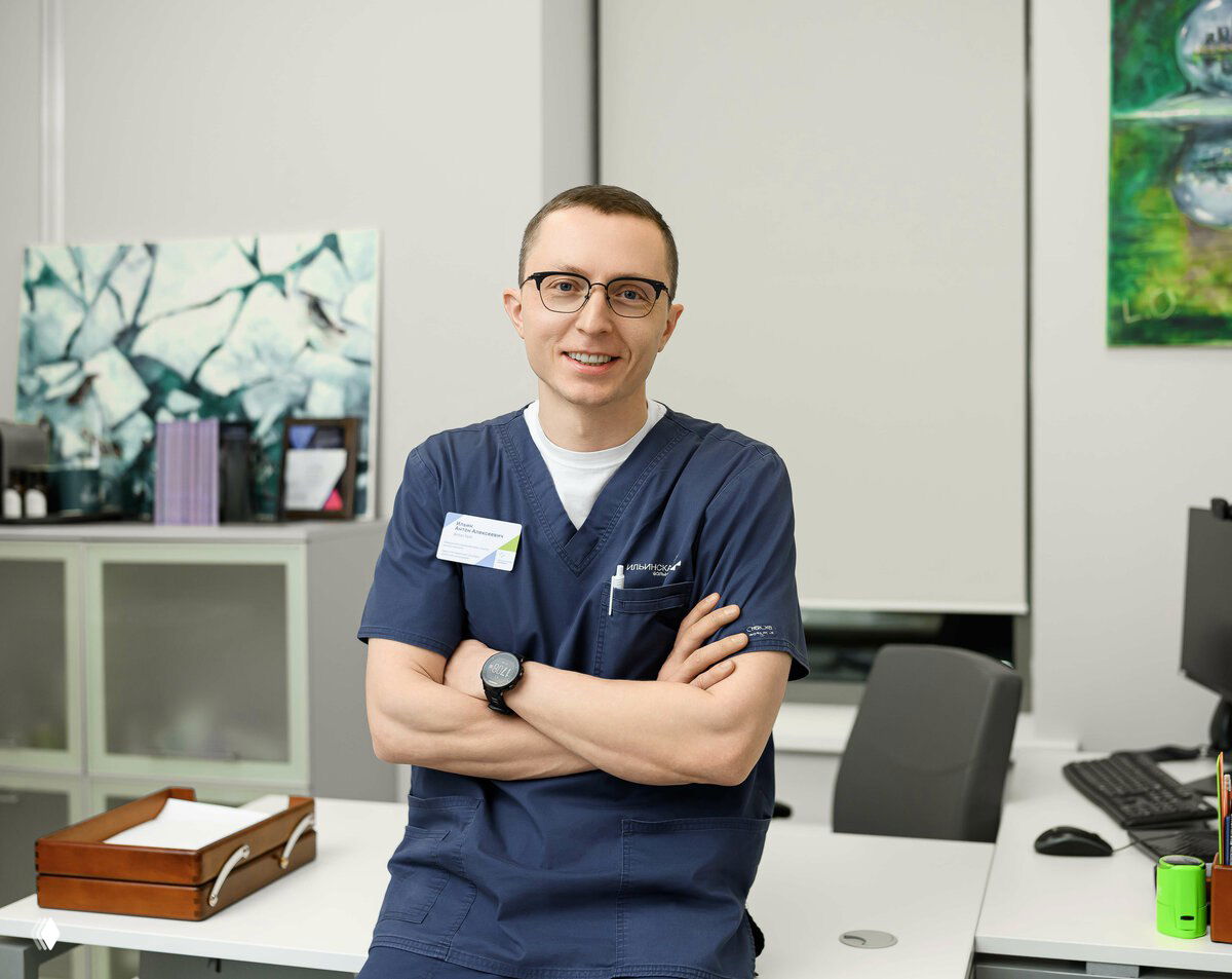 Молодой врач в тёмно-синей медицинской форме стоит в кабинете с скрещёнными руками и улыбается, на столе видны документы и рабочая зона.