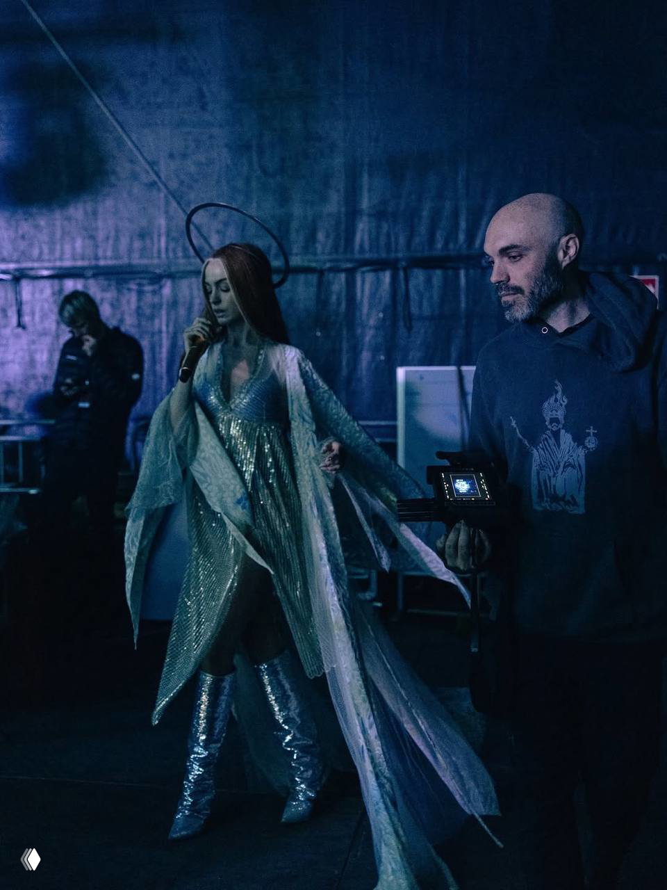 Энн Хэтэуэй в длинном переливающемся костюме и высоких сапогах на сцене; оператор и съёмочное оборудование видны рядом, атмосферный кадр