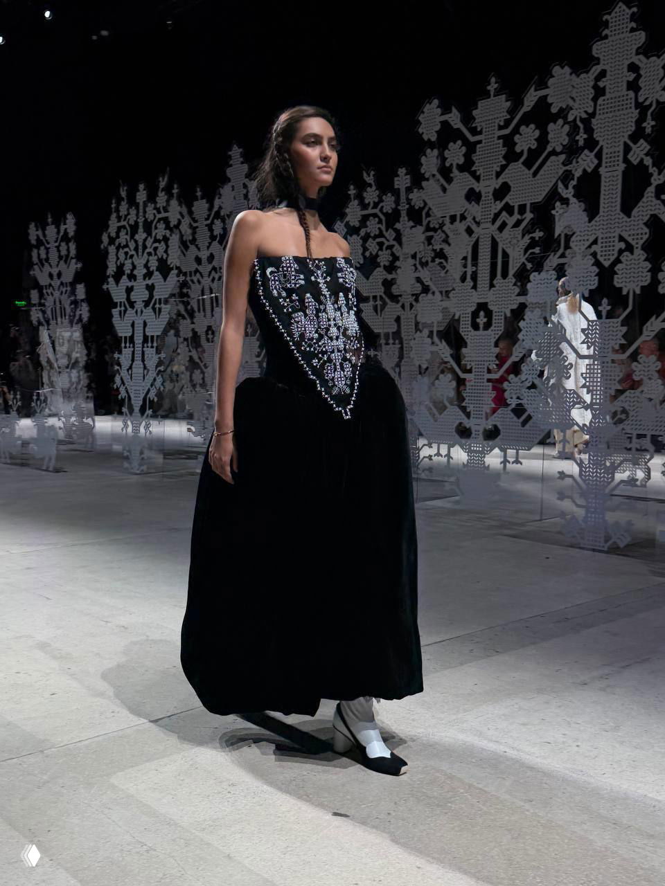 Модель в чёрном платье с вышитым корсетом шагает по подиуму на фоне белых ажурных декораций и световых прожекторов показа.