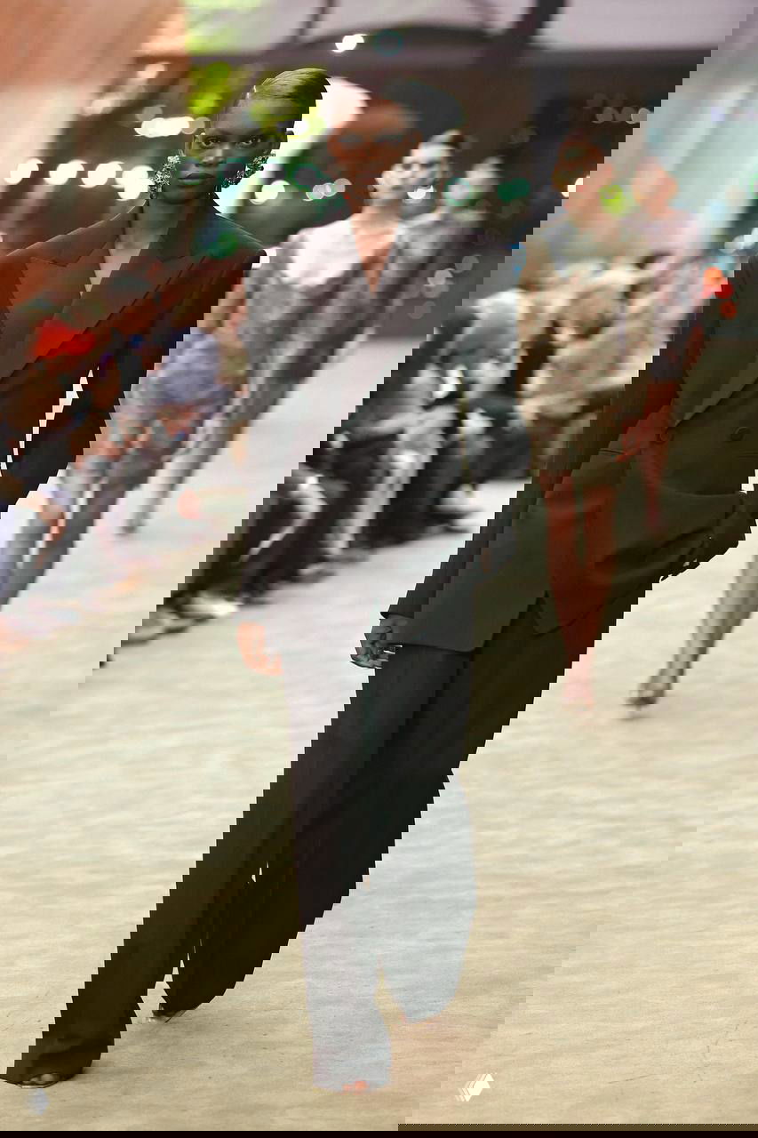 Модель в чёрном двубортном костюме шагает по подиуму Saint Laurent; за ней видны другие модели и зрители показа.