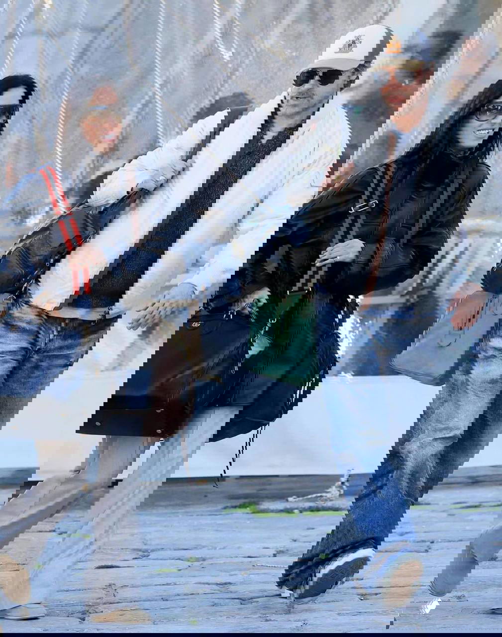 Папарацци-снимок: мужчина и женщина в повседневной одежде идут по каменной набережной с крупными дорожными сумками на фоне уличной сцены и людей.