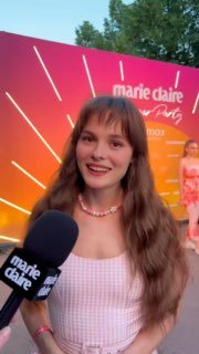 Улыбающаяся женщина на вечернем мероприятии Marie Claire, даёт интервью с микрофоном на фоне яркого брендового баннера.