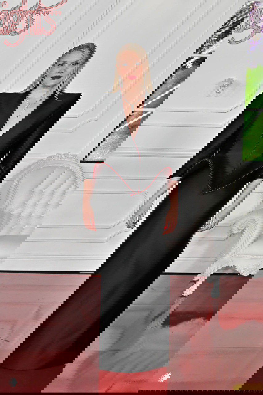 Женщина в строгом чёрном платье с подчеркнутыми плечами и глубоким декольте позирует на фоне белого дивана и элементов интерьера премьеры.