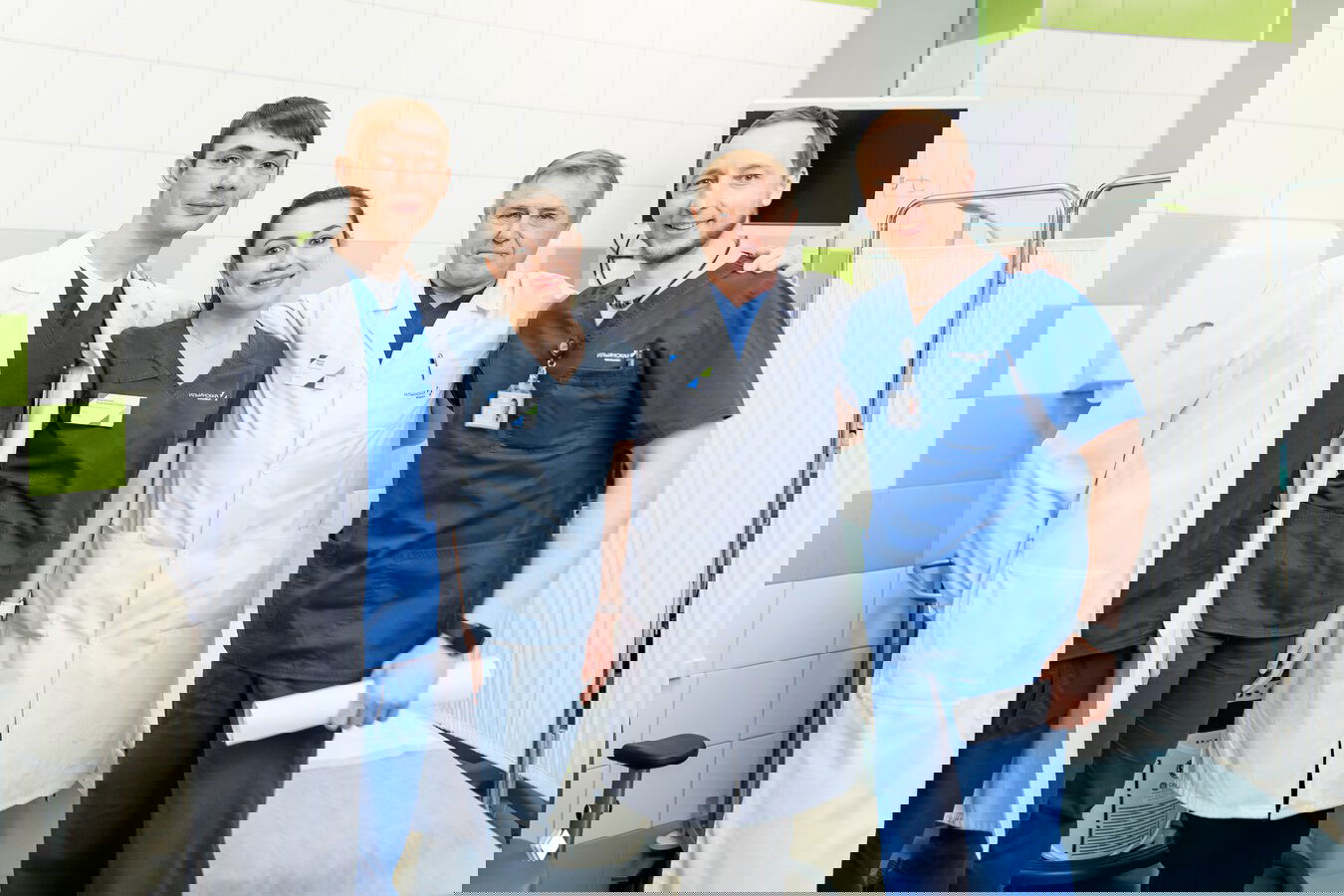 Группа из четырёх медработников в халатах и медицинской форме позирует в процедурном кабинете клиники на фоне оборудования и перегородок.