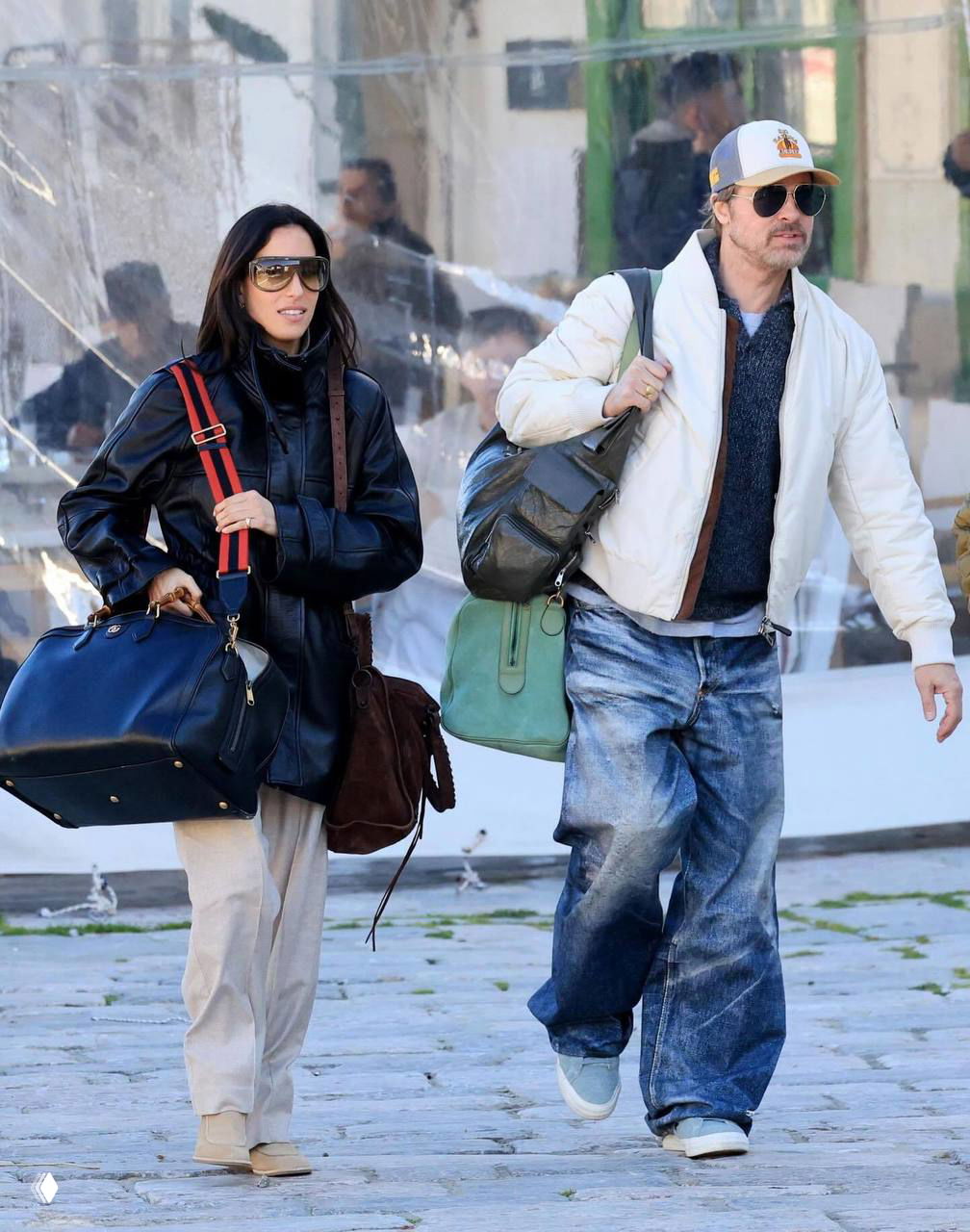 Папарацци-фото: пара идёт по мощёной улице с дорожными сумками, повседневные пальто и джинсы, вокруг — уличная обстановка и люди.