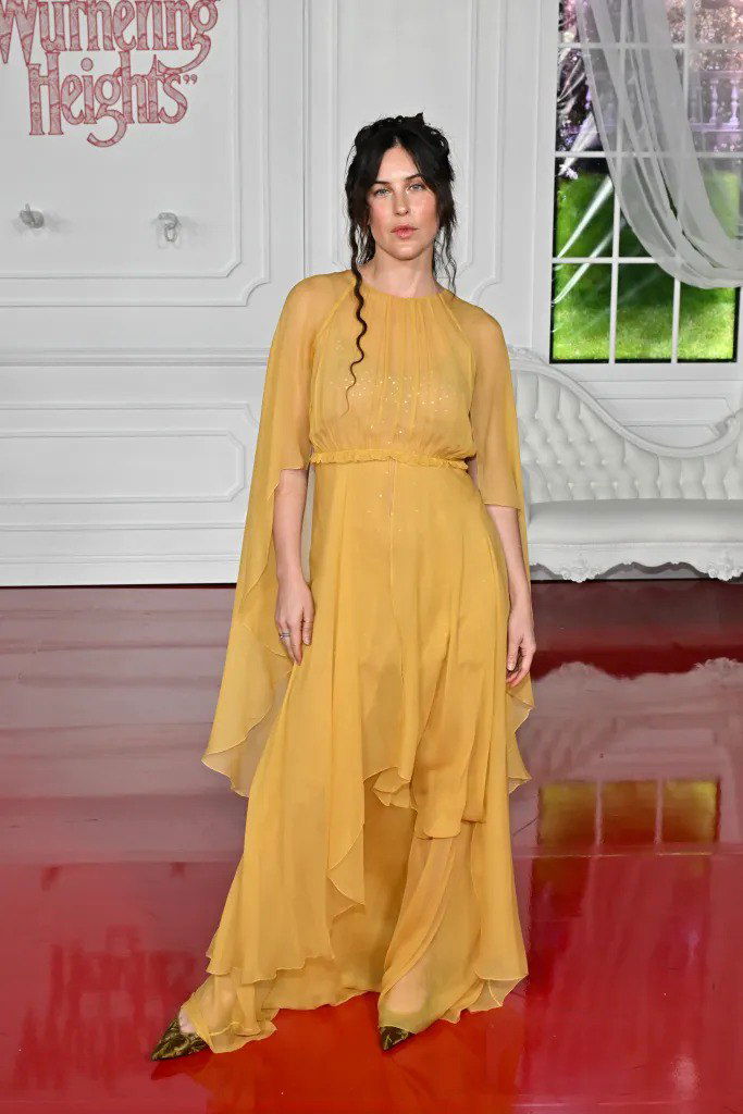 Гостья в лёгком жёлтом струящемся платье идёт по красной глянцевой дорожке у декоративного интерьера и оконного проёма — романтичный образ.