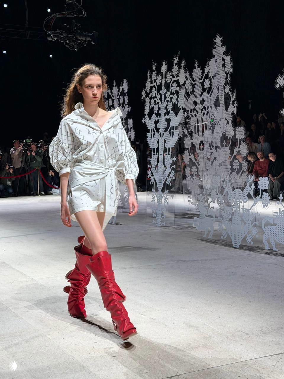 Модель в лёгком белом платье и ярко‑красных сапогах идёт по сцене; контрастная обувь подчёркивает ретро‑мотивы коллекции «Приданое».