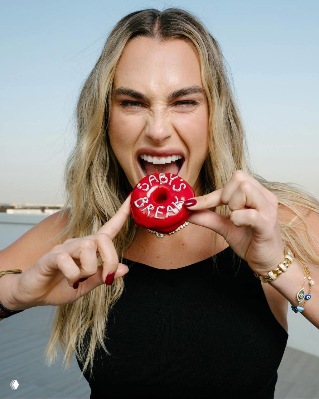 Крупный портрет Арины Соболенко, она держит красный пончик с надписью и делает игривое выражение лица на фоне неба в Дубае.