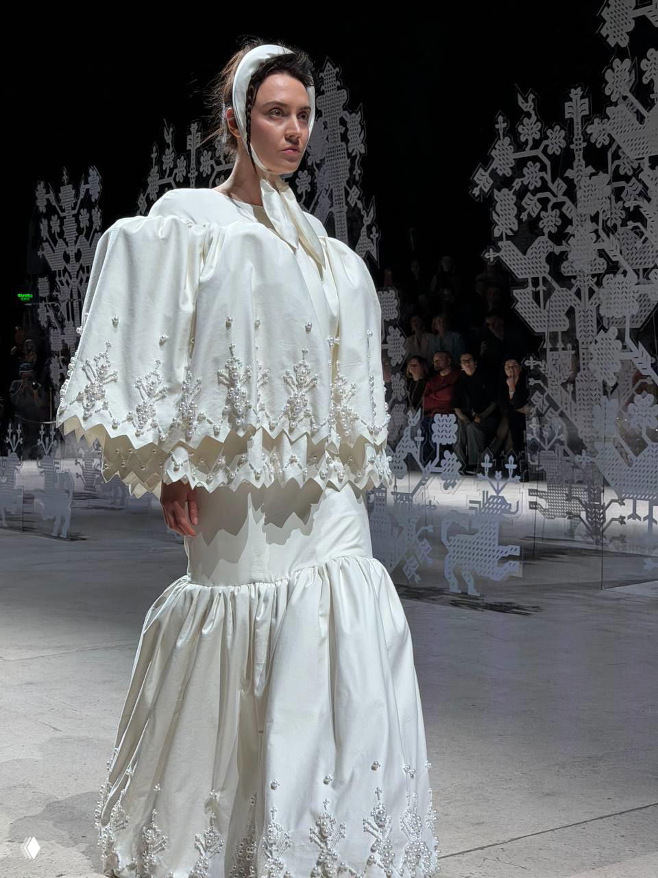 Модель в белом наряде с объёмными рукавами и украшенной юбкой проходит мимо ажурных резных декораций на показе бренда.