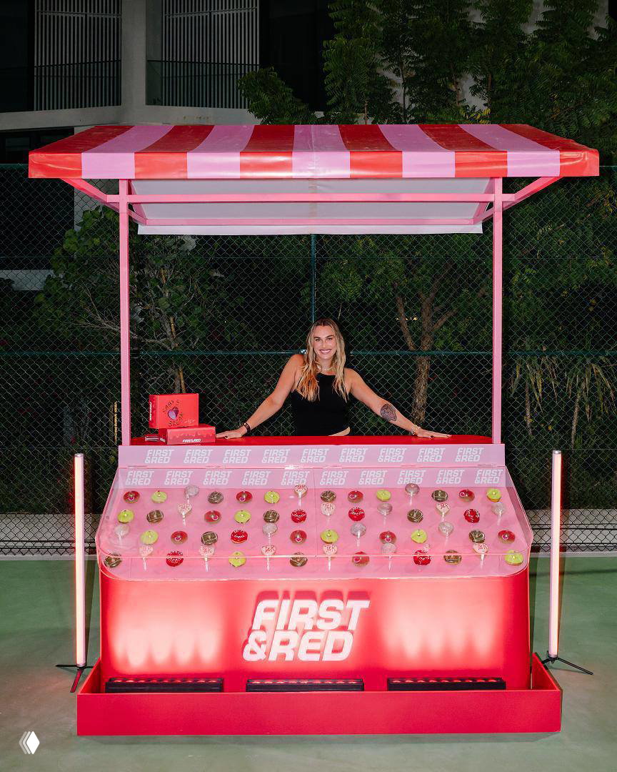 Арина Соболенко стоит за розовым стендом First&Red с рядом пончиков, вечернее освещение, сетка корта и деревья на заднем фоне.
