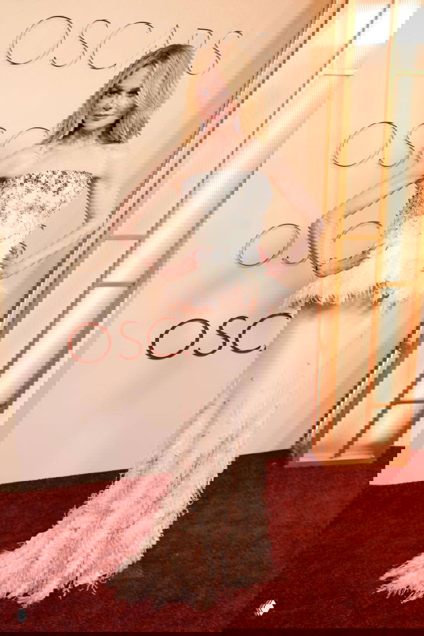 Женщина в утончённом светло-розовом платье с перьевыми и блестящими деталями на фоне бежевой фотостены Оскара 2026.