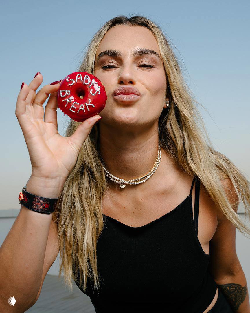 Крупный план: Арина Соболенко держит красный пончик у губ, делает вид поцелуя, на ней жемчужное ожерелье и лёгкий макияж.