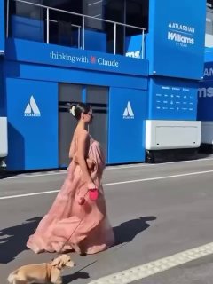 Кадр из видео: Александра Леклер в розовом платье идет по дорожке паддока, рядом на поводке маленькая собака, синие конструкции на фоне.